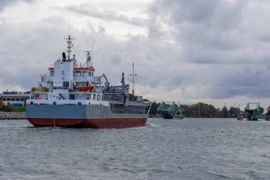 Orta ölçekli bir kargo gemisi, kıç tarafı izleyiciye dönük bir şekilde bir kanaldan geçiyor, açık yeşil salıncak köprüsünü ve kıyıdaki sanayi binalarını manevra yaparak geçiyor..