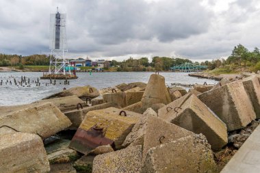 Su kanalı sahnesinde büyük beton bloklar, beyaz bir navigasyon kulesi ve uzak mesafede karanlık, dramatik bulutlu bir gökyüzüne karşı kurulmuş eski yeşil bir köprü var..