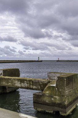 Eski bir rıhtımın çürüyen beton yapısı dalgalı suyun üzerine çöker, uzak liman girişini, ağır, bulutlu bir gökyüzünün altında yeşil ve kırmızı navigasyon fenerleriyle çerçeveler..