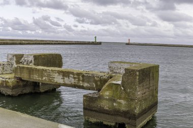 Çürüyen beton iskele yapısının geniş görüşü suya açılıyor. Liman kanalı girişi, uzun dalgalar ve renkli navigasyon fenerleri var..