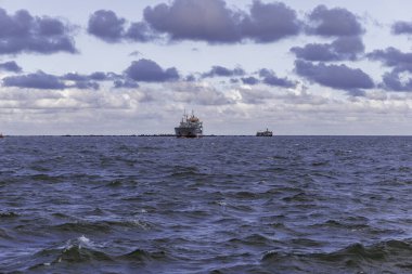 Bir kargo gemisi açık bir şekilde dalgalı denizde yol alırken, dalgalı bir dalgakıran ve dramatik, mor renkli bulutların hakim olduğu bir gökyüzü tarafından çerçevelenir..