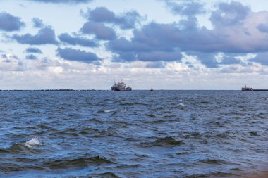 Bir kargo gemisi ve küçük bir römorkör liman girişine doğru yol alıyor. Dalgalı, karanlık sularda, dramatik bulutlu bir gökyüzünün altında..