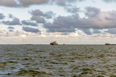 Dalgalı açık denizde bir kargo gemisi ve küçük bir römorkör, dalgalı bir açık denizde, dalgalı bir ufuk ve dramatik, kara bulutlar tarafından çerçevelenmiş.