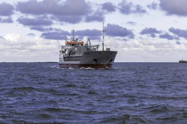 Dalgalı denizde bir kargo gemisi yelken açar, uzak bir liman girişine yaklaşır. Dalgakıran bir dalgayla işaretlenir, mor renkli, dramatik bulutlu bir gökyüzünün altında..