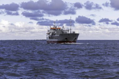 Orta büyüklükteki bir kargo gemisi, beyaz ve mor renkli bulutları olan dinamik bir gökyüzüne karşı karanlık ve dalgalı sularda izleyiciye doğru yol alır..