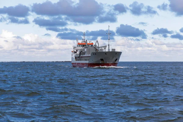 Orta büyüklükteki bir kargo gemisi, beyaz ve mor renkli bulutları olan dinamik bir gökyüzüne karşı karanlık ve dalgalı sularda izleyiciye doğru yol alır..