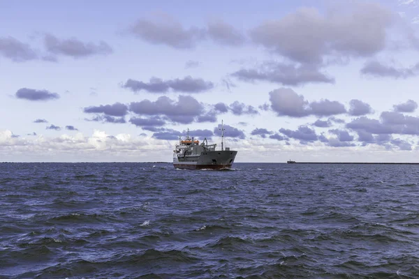 Açık denizde yelken açan bir kargo gemisinin uzaktan görünüşü, sağda dalgalı bir yapıyla çevrili, geniş bir gökyüzünün altında, dağınık, mor renkli bulutlarla..