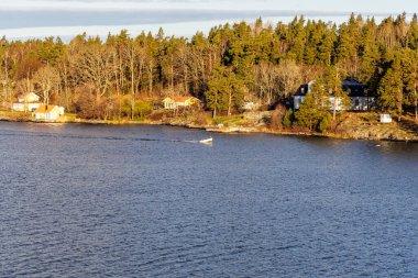 Sarı ve beyaz evleri olan engebeli, ormanlık bir kıyı şeridinin orta manzarası ve yanından geçen bir motorlu tekne, kıyı ikametgahını, seyahatini ve kanalın faaliyetlerini vurguluyor..
