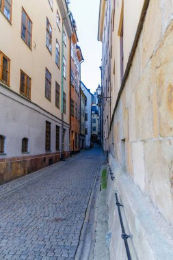 Dar, yüksek duvarlı bir kaldırım taşı sokak manzarası. Uzun, pastel renkli binalar arasında sokağın keskin bir şekilde kaybolduğu yer. Taş temeli ve tarihi feneri olan..