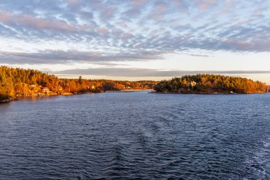 Takımadalardaki ormanlık adaların geniş panoramik manzarası, sabahın erken saatlerinde ya da öğleden sonra güneş ışığı kıyı ağaçlarını ve evleri aydınlatırken.