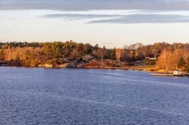 Huzurlu bir körfez manzarası. Ormanlı, kayalık kıyıları ve küçük bir deniz kenarı binası. Sakin, arzulanan bir konumu temsil ediyor..