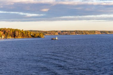 Yoğun, alçak, ormanlık bir kıyı şeridiyle çevrili, kayalık bir adada küçük beyaz bir seyir feneri olan sakin, karanlık bir su kanalı geniş bir açıdan görülüyor..