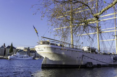 Stockholm 'de demirlemiş tarihi AF Chapman gemisinin etkileyici yayı. Eski şehir binaları ve güneşli arka planda görünen modern bir yat..