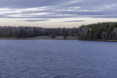 Alacakaranlıkta dramatik bulutlu bir gökyüzünün altında, gölde ya da deniz kıyısında karışık bir ormana kurulmuş, uzak bir İskandinav ahşap kulübesinin manzarası..