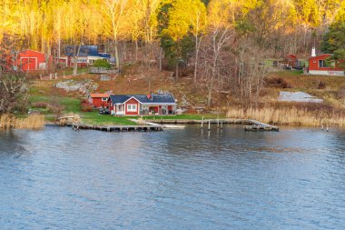 Kayalık ve kıyı şeridinde kayalık rıhtımları olan geleneksel kırmızı ve beyaz ahşap kulübelerin yakın görüntüsü, altın saat güneş ışığıyla yıkanmış..