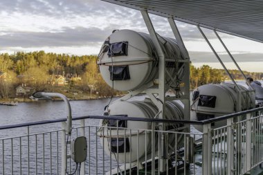 İstiflenmiş, silindirik, beyaz cankurtaran botları arka planda İskandinav kıyı şeridi manzaralı bir feribot veya yolcu gemisinin güvertesinde emniyete alındı..