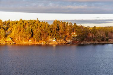 Sıcak güneş ışığı altında küçük beyaz ve yeşil bir seyrüsefer fenerinin yer aldığı kayalık, ormanlık bir sahil şeridinde gözlerden uzak, gözlerden uzak bir ev manzarası..