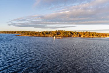 Engebeli, ormanlı bir takımada su kanalına geniş bir perspektif, dramatik bir gökyüzü altında küçük beyaz bir işaret feneri, ölçeği ve yolculuğu vurguluyor.