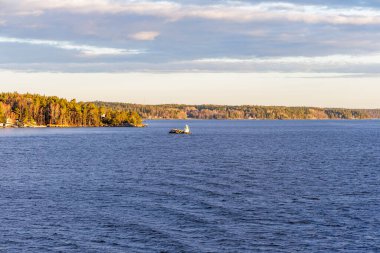 Geniş bir deniz kanalı manzarası, kayalık tabanındaki küçük beyaz bir feneri bulutlu bir gökyüzünün altındaki geniş, ormanlık İskandinav kıyı şeridinin arka planına vurguluyor..