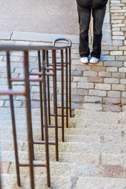 Dik bir taş merdivenin üstünden yüksek açılı bir atış. Ön planda koyu metal bir korkulukla kaldırım taşının üstünde duran birinin alt bacaklarını gösteriyor..