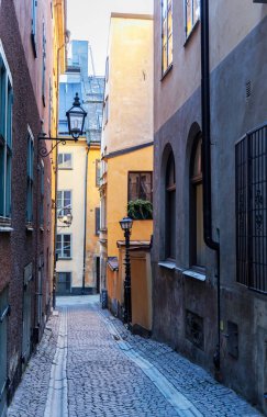 Avrupa 'nın tarihi bir şehrinde (Gamla Stan) dar, sessiz bir kaldırım yolu manzarası, duvarına antik siyah bir fener monte edilmiş uzun, renkli binalarla çevrili..