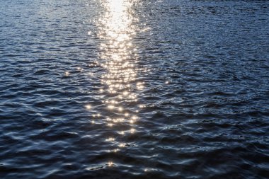 Belirgin, derin dalgalanmaları ve güçlü, çarpık ışık kaynağının yansımasını içeren çok koyu mavi su yüzeyinin sıkı, tam kare görüntüsü yüksek kontrastlı soyut bir doku oluşturur..