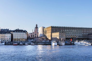 Stockholm Kraliyet Sarayı 'nın ve Gamla Stan' in yakınındaki rıhtım bölgesinin manzarası. Açık bir gökyüzünün altındaki rıhtım boyunca demirlemiş tekneleri ve tarihi binaları var..