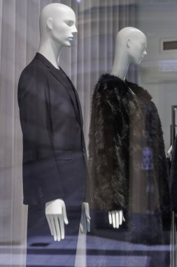 Bir vitrinde sergilenen iki beyaz mankenin yan görüntüsü: koyu takım elbiseli bir erkek manken ve koyu renk kürklü bir kadın model..