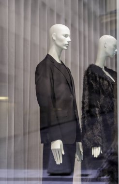 Bir dükkan vitrininde iki beyaz manken: biri siyah takım elbiseli, diğeri siyah kürklü, beyaz perde arkasında..