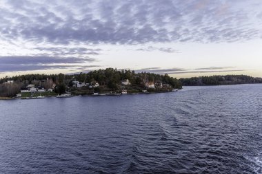 Tipik İskandinav ahşap evleri ve tekne rıhtımlarının bulunduğu ormanlık ada kıyı şeridi manzarası bulutlu bir akşam veya gün batımında bir yolcu gemisinden görülüyor..