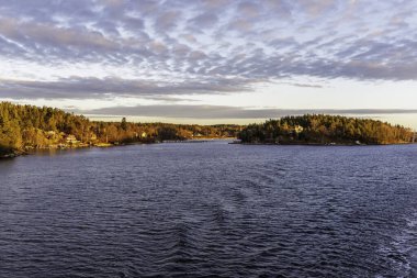 Takımadalardaki ormanlık adaların geniş panoramik manzarası, sabahın erken saatlerinde ya da öğleden sonra güneş ışığı kıyı ağaçlarını ve evleri aydınlatırken.