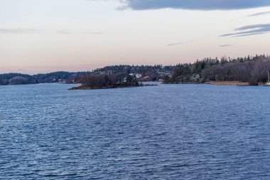 Geniş açılı deniz manzaralı, dalgalı karanlık suyla uzak arka planda dağınık beyaz ve kırmızı evleri olan bir orman adasına doğru..