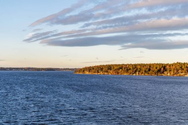 Sağdaki alçak, ormanlık bir kıyıya doğru geniş bir kanal boyunca geniş bir manzara çizgili bulutlarla dramatik bir gökyüzünün altında geniş bir açık deniz geçidine işaret ediyor..