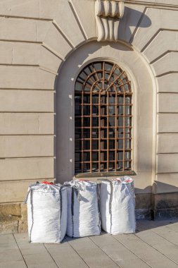 Önünde üç büyük, beyaz, örülmüş inşaat torbası bulunan, süslü kemerli pencereli ve paslı demir parmaklıklı klasik bir taş cephenin yakın çekim, dikey çekimi..