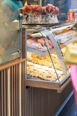 Gıda tezgahında cam bir vitrin, üst rafta parlak kırmızı elma şekerli, birden fazla renkli jöleli ve serpilmiş çörekleri gösteriyor..