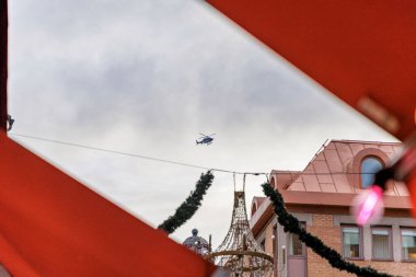 Bir helikopter bulutlu bir gökyüzüne doğru uçuyor, bir tente tarafından çerçevelenmiş ve ön planda bir binanın çatısı ve askıya alınmış şenlikli çelenkler bulunuyor..