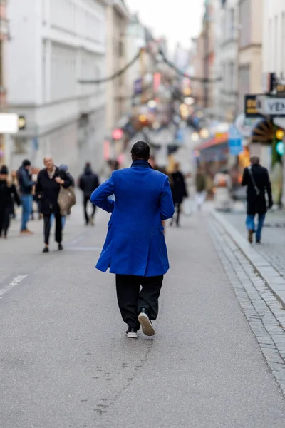 Arka planda, parlak, mor, diz boyu bir ceket giyen bir adam var. Şehrin yaya caddesinde yürüyor. İnsanların bulanık bir geçmişi ve tatil süslemeleri var..