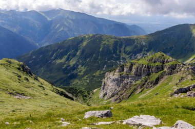 Dik yeşil yamaçları ve beyaz bulutların altında kayalık oluşumları olan derin dağ vadisi. Çevre güzelliğinin, biyolojik çeşitliliğin ve yaz yürüyüşü rotalarının altını çizmek.