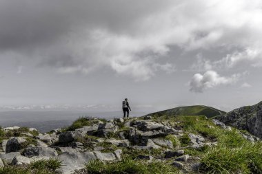 Sırt çantalı bir insan dramatik bulutlu bir gökyüzünün altında geniş bir dağ manzarasına bakar. Liderliğin, kişisel gelişimin ve açık hava macera turizminin sembolü.