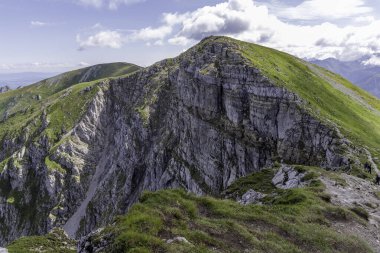 Dramatic view of vertical limestone cliffs and green mountain slopes overlooking a distant valley. Focus on geology, natural wonders, and high-altitude tourism.