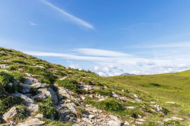 Ön planda kayalık bir dağ sırtının alçak açılı görüntüsü gözü yumuşak yeşil bir yamaca ve ince bir gökyüzünün altındaki uzak bir zirveye yönlendiriyor..