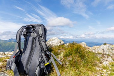 Yaz yürüyüşü sırasında uzak mavi tepeler ve beyaz bulutlara karşı kayalık bir dağın zirvesinde dinlenen siyah bir sırt çantası..