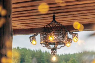 Terasta ahşap bir tavandan sarkan parlayan ampulleri olan antika tarzı büyük bir metal avize. Konukseverlik, lüks etkinlikler ve ışıklandırma tasarımı için ideal.