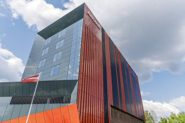 Kırmızı yüzgeçli, camdan pencereli ve Letonya ulusal bayrağının rüzgarda dalgalandığı modern mimari yapının yukarı açısı.