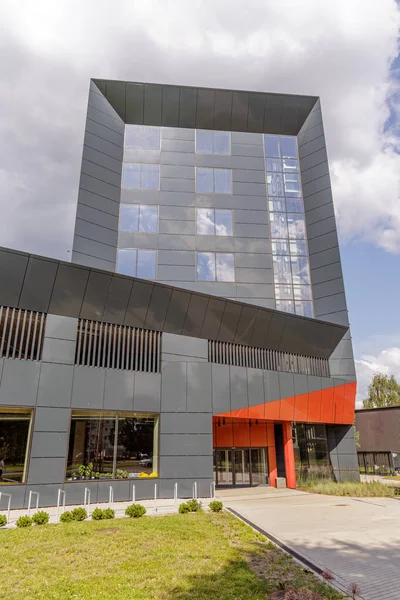 Geniş açılı, koyu gri kaplamalı, girişte turuncu aksanlı ve gökyüzünü yansıtan büyük camlı modern bir bina..