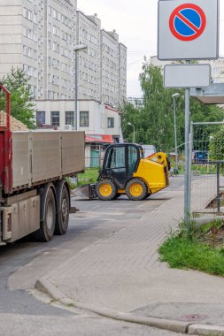 Küçük sarı bir fren dümeni yükleyicisi şehir binalarının yakınındaki bir caddeye park etmiş. Şehir altyapısının gelişimini ve yol çalışmalarını gösteriyor..