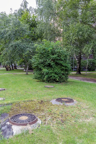 İkamet binaları ve ağaçların yakınındaki yeşil çimlerde bulunan metal rögar kapakları kentsel altyapı ve bakımı temsil ediyor..