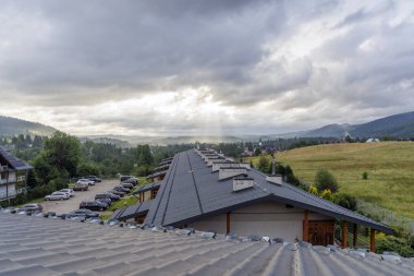 Dağlık bir tatil köyü binası ve vadide ağır bulutları delip geçen güneş ışınlarıyla dolu yeşil bir tarla..