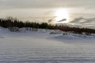 Güneş, karlı bir kıyı manzarasının üzerinde kum tepeleri ve çalılarla birlikte bir bulut katmanının arkasında batar. Sıcak altın ışık, beyaz kara huzurlu bir doğa görüntüsüyle vurur..