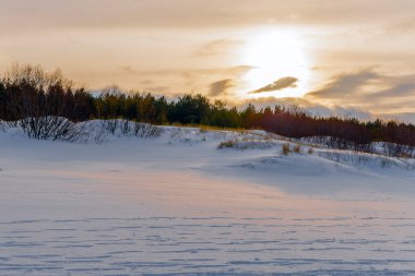 Güneş, karlı bir kıyı manzarasının üzerinde kum tepeleri ve çalılarla birlikte bir bulut katmanının arkasında batar. Sıcak altın ışık, beyaz kara huzurlu bir doğa görüntüsüyle vurur..